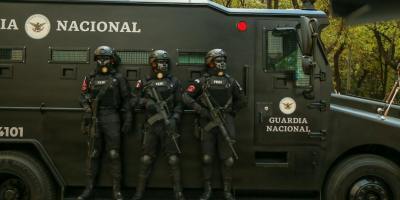 Three armed Guardia Nacional officers in black tactical gear stand shoulder-to-shoulder beside a matte-black armored vehicle on a tree-lined city street.