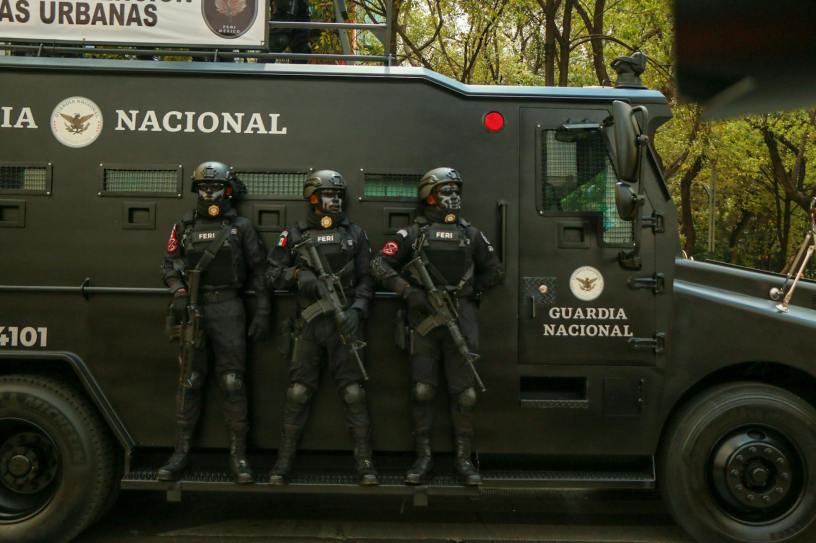 Three armed Guardia Nacional officers in black tactical gear stand shoulder-to-shoulder beside a matte-black armored vehicle on a tree-lined city street.