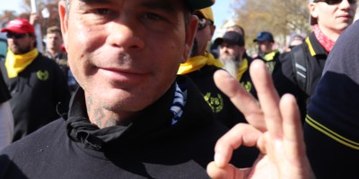 Close-up of a Proud Boys member at the Million MAGA March making the “OK” hand sign, wearing branded clothing and surrounded by other members in coordinated yellow and black attire.