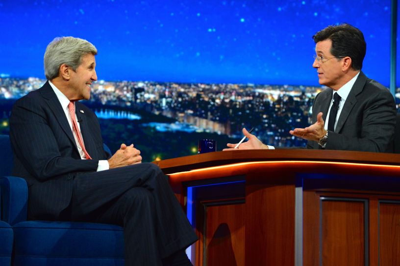 U.S. Secretary of State John Kerry in conversation with Stephen Colbert on The Late Show, seated across from each other at a studio desk with a city skyline in the background.