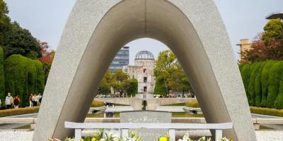 View through the Hiroshima cenotaph arch toward the Atomic Bomb Dome, with flowers laid at the memorial and trees lining the peaceful park walkway.