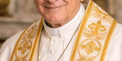 Pope Benedict XVI wearing white papal vestments with gold embroidery and a gold cross, standing in a Vatican interior setting.