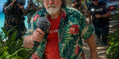 A bearded field reporter in a tropical shirt walks toward the camera holding a microphone while police and photographers follow behind him on a palm-lined beach with flashing lights in the background.