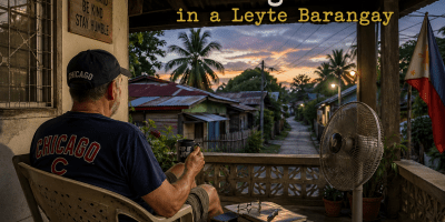 American man wearing a Chicago Cubs shirt sitting on a porch in a Leyte barangay, Philippines, at dusk with a quiet rural street in the background