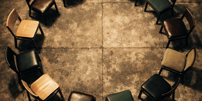 Circle of twelve empty chairs in various colors on concrete floor