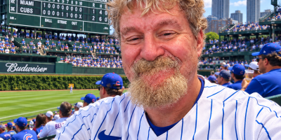 AI-created image of WPS News Editor-in-Chief Cliff Potts wearing a Chicago Cubs jersey and sitting among fans in the Wrigley Field bleachers, celebrating the Cubs clinching a 2026 postseason berth.